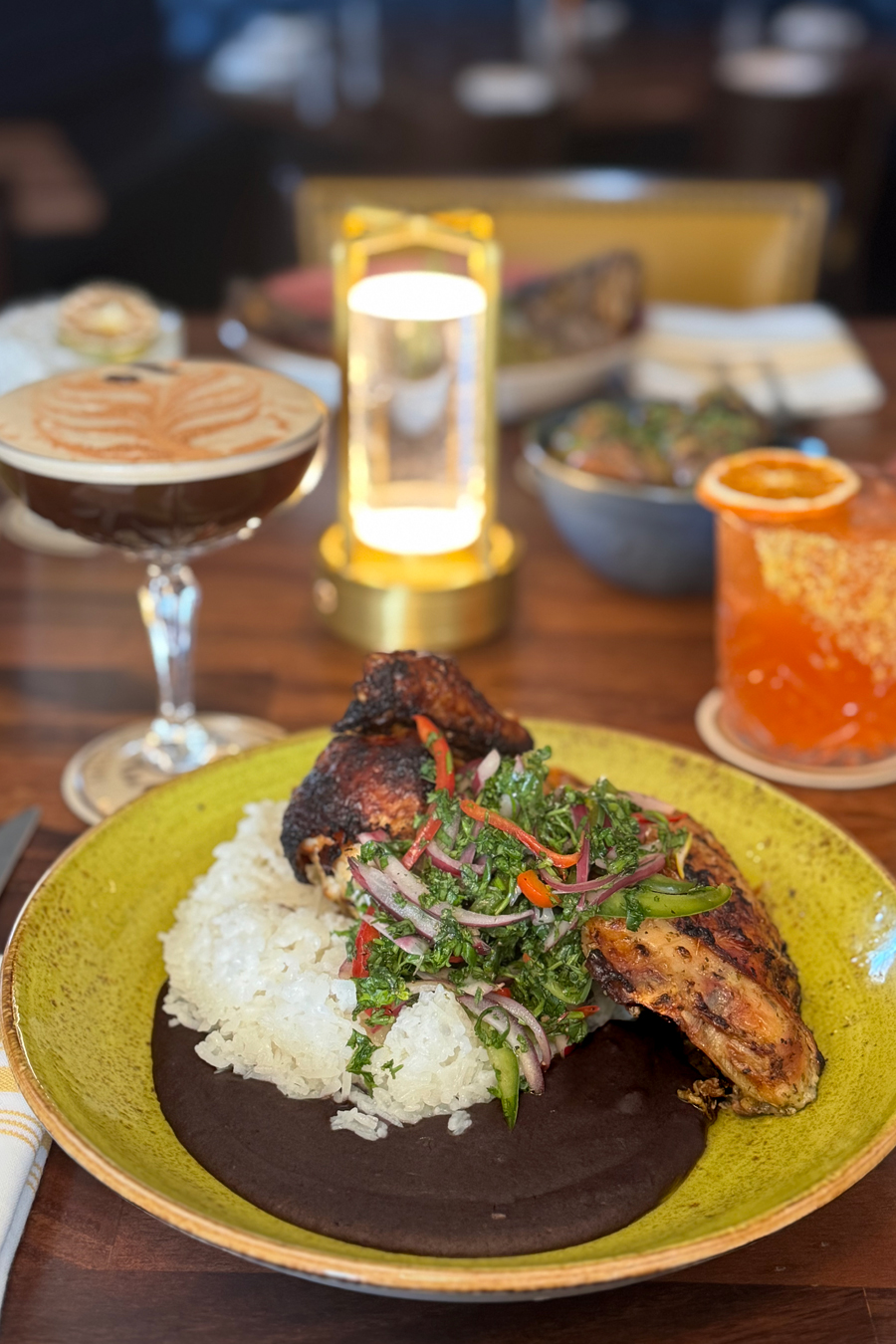 A vibrant dish featuring grilled chicken over rice, topped with fresh herbs and vegetables on a green plate. Nearby are a cocktail, an orange drink, and a lit lantern, conveying elegance and warmth.